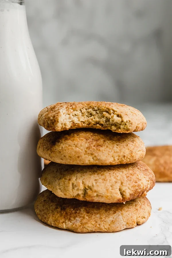A perfectly stacked trio of pumpkin snickerdoodle cookies, displaying their sugary coating, placed beside a refreshing glass of plant-based milk on a light surface.