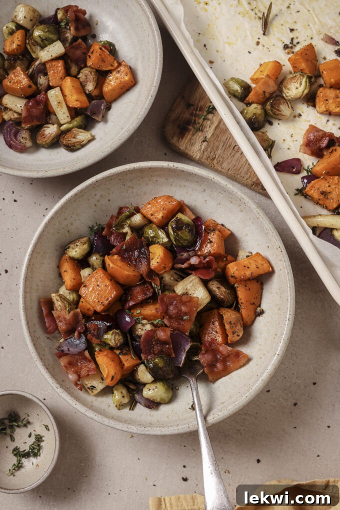 A vibrant bowl of roasted harvest vegetable hash, rich with sweet potato, Brussels sprouts, parsnips, and carrots, topped with crispy bacon, with a baking sheet in the background