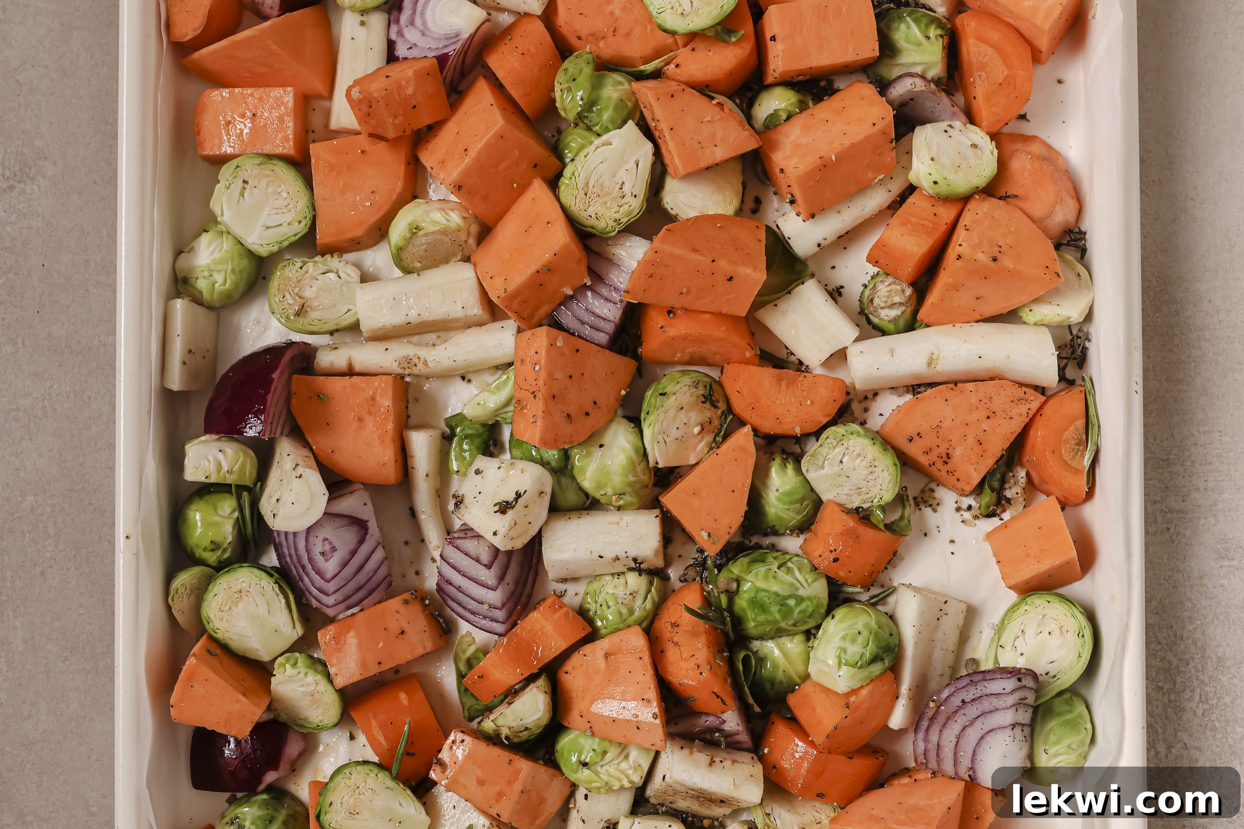 Chopped sweet potato, parsnip, Brussels sprouts, and red onion are spread evenly on a baking sheet, seasoned and ready for roasting.