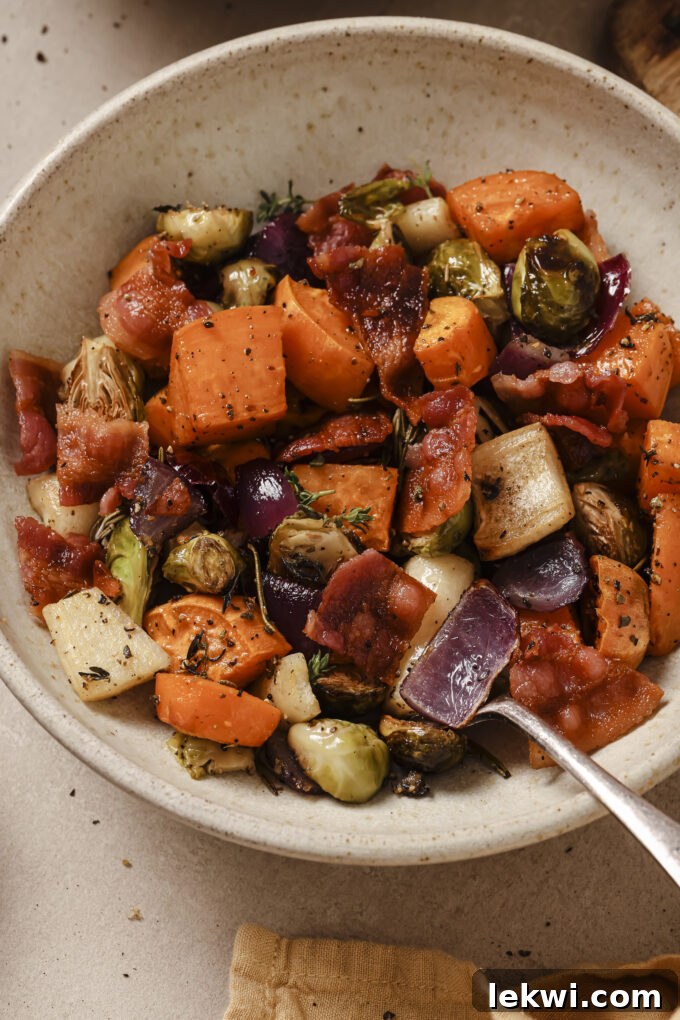A close-up of a bowl filled with roasted harvest vegetable hash, featuring golden sweet potatoes, parsnips, Brussels sprouts, red onion, and savory bacon pieces, garnished with fresh herbs.