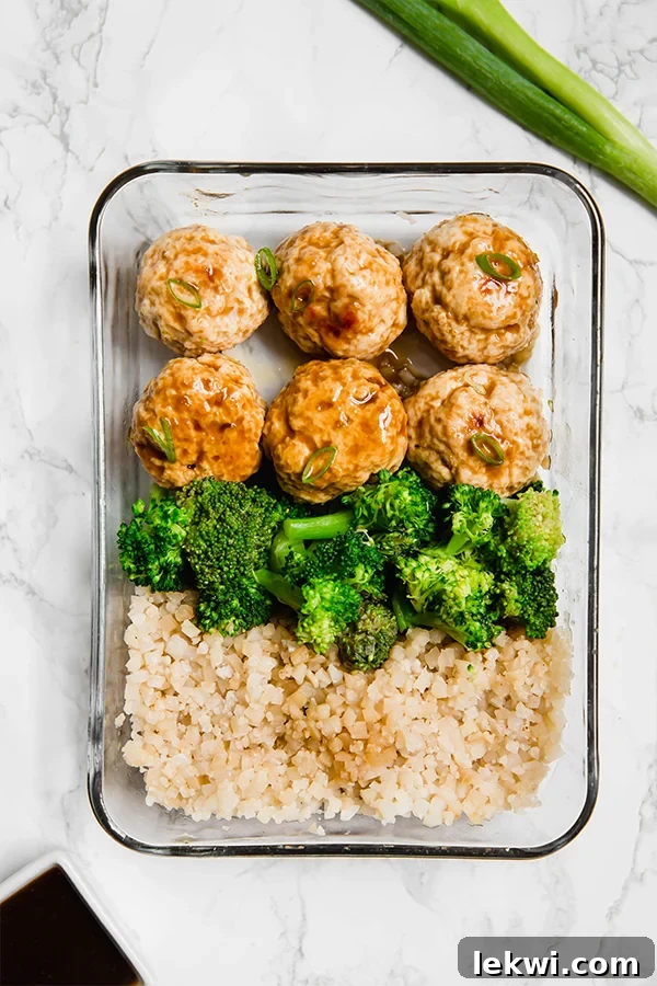 A close-up of a single meal prep container filled with tender teriyaki chicken meatballs, vibrant broccoli florets, and fluffy cauliflower rice.