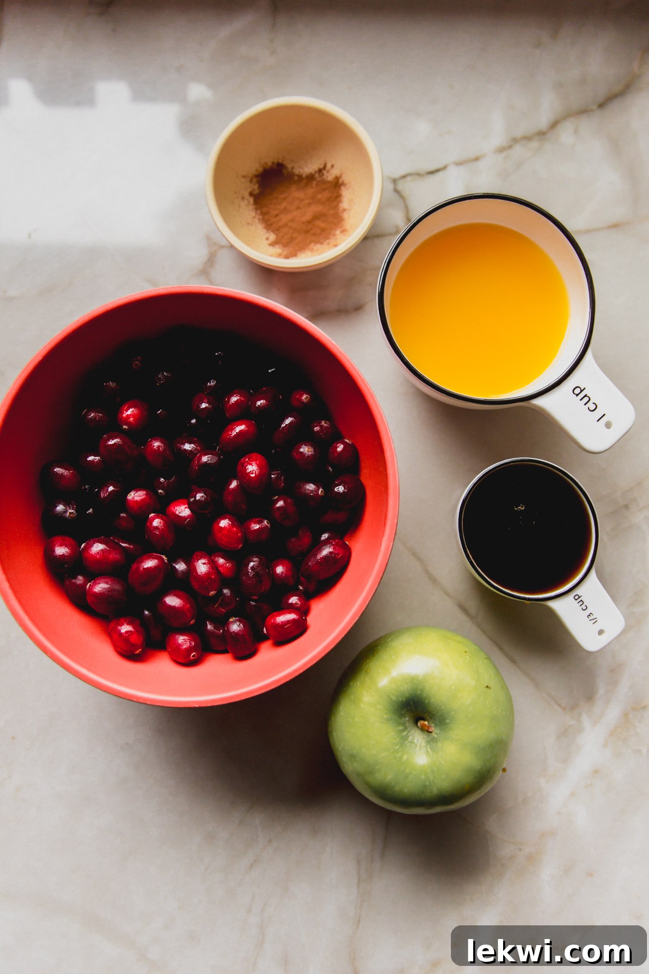 Separated ingredients for apple cranberry sauce: fresh cranberries, a diced Granny Smith apple, maple syrup, orange juice, cinnamon, and orange zest.