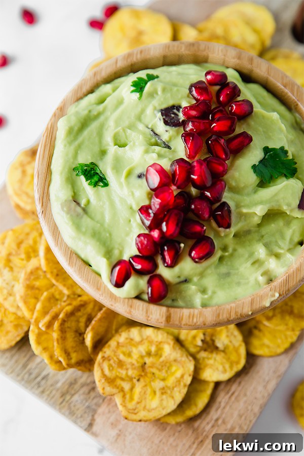 A rustic wooden bowl brimming with fresh pomegranate guacamole, surrounded by crisp plantain chips for dipping.