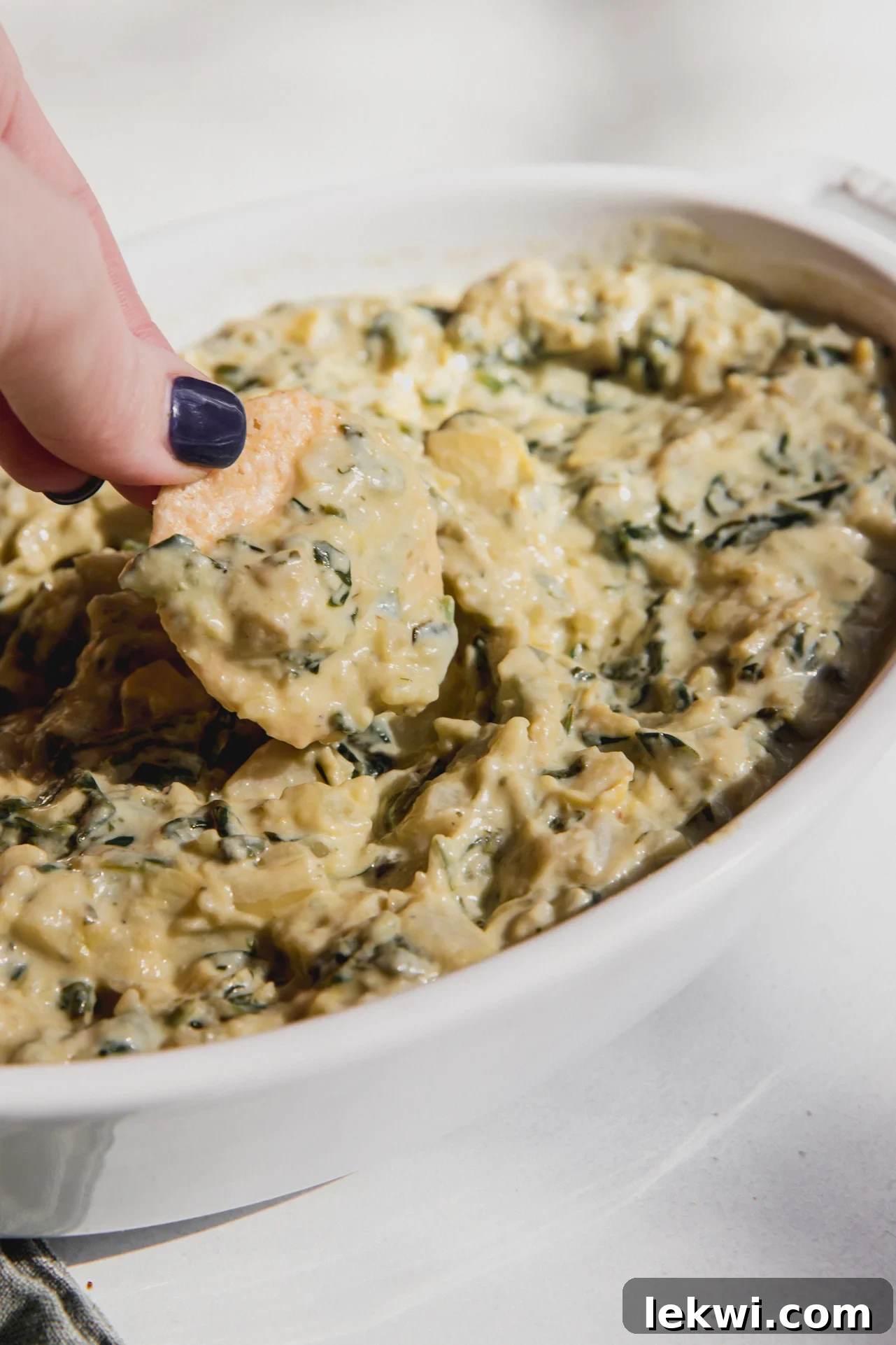 A cracker dipping into spinach and artichoke dip in a white bowl, showcasing the creamy texture.
