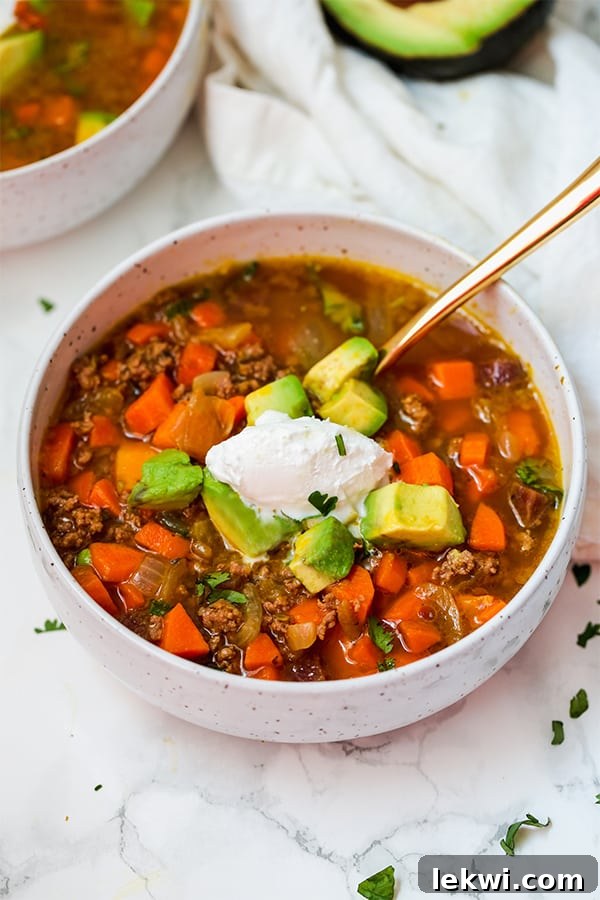 A white bowl of vibrant Paleo & AIP Pumpkin Chili, topped generously with a swirl of coconut cream and perfectly sliced avocado, resting on a rustic wooden table.
