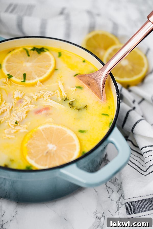 A rustic blue Dutch oven brimming with aromatic Greek Lemon Chicken Soup, with a silver serving spoon gently stirring the rich, golden broth and tender chicken pieces.