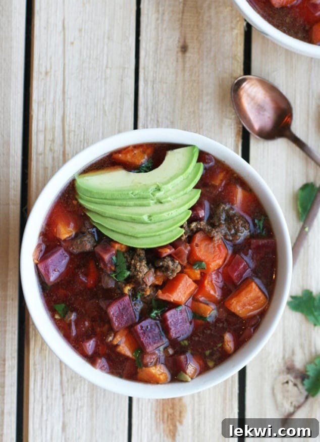 A rustic wooden table features a white ceramic bowl filled with vibrant AIP Chili, generously topped with perfectly sliced avocado and fresh cilantro, alongside a small lime wedge.