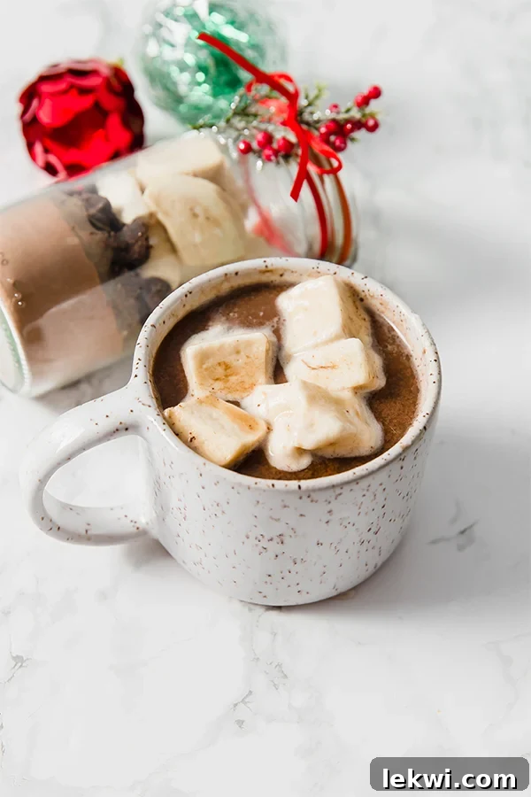 A ceramic mug filled with hot coco and toped with marshmallows next to a glass container of diy cocoa.