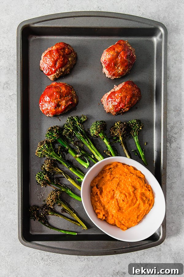 A sheet pan loaded with perfectly roasted broccolini, golden mini meatloaves, and a small white bowl of creamy mashed sweet potatoes.