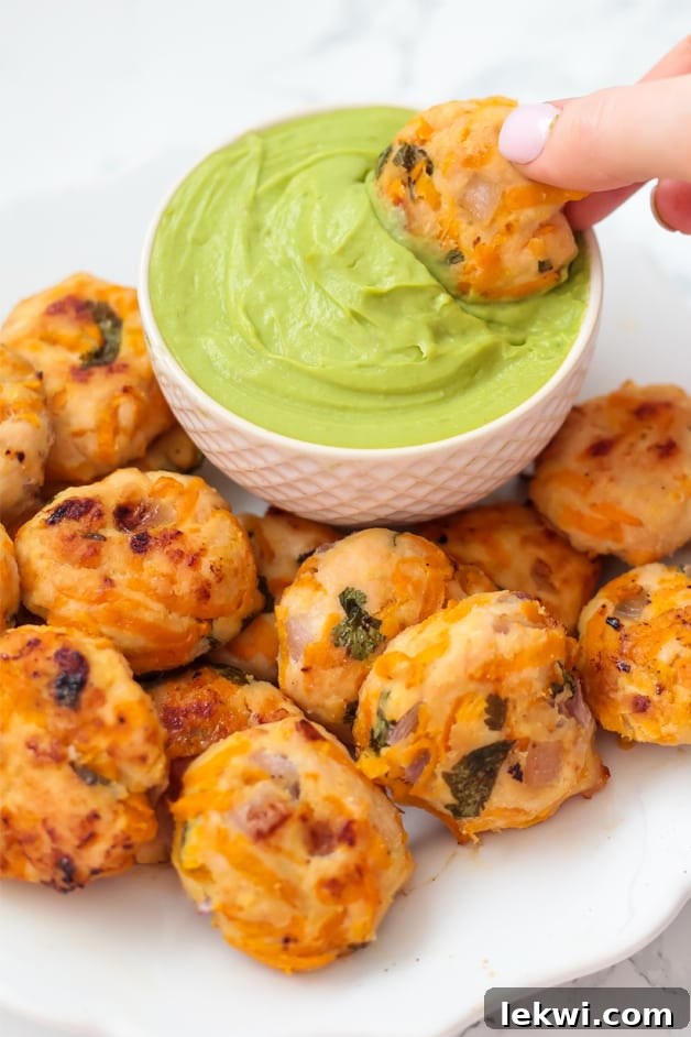 Many savory Mexican chicken poppers artfully arranged around a small white cup filled with creamy guacamole, with one chicken popper being dipped into it.