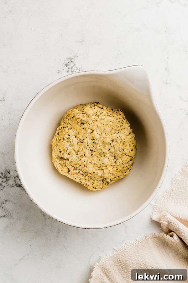 A close-up view of the cauliflower breadstick dough, freshly mixed and ready to be shaped, showcasing its pliable texture and flecks of herbs, in a mixing bowl.