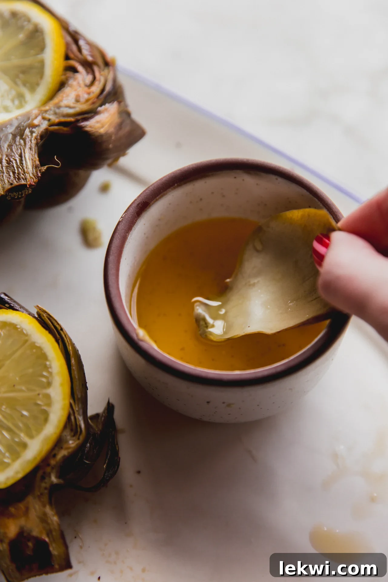 A perfectly baked artichoke leaf being dipped into a small bowl of melted ghee, highlighting the delicious eating experience.