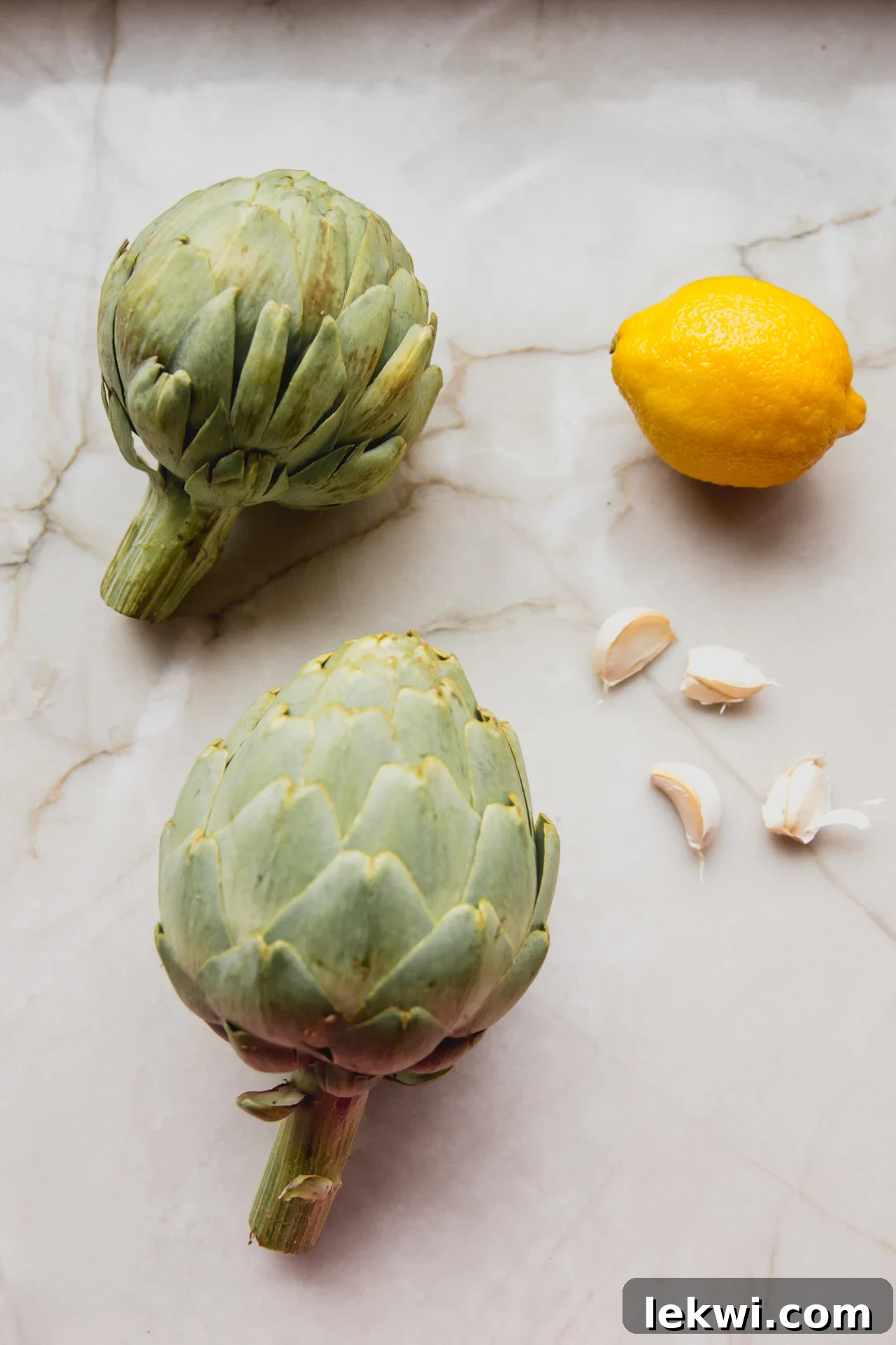 Two fresh artichokes, a bright yellow lemon, and several garlic cloves neatly arranged on a white marble countertop, ready for preparation.