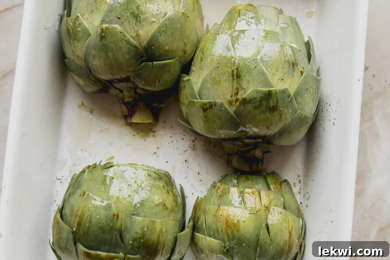 Four halved artichokes arranged in a baking dish, leaf-side up and ready for the oven, with garlic and seasonings visible.