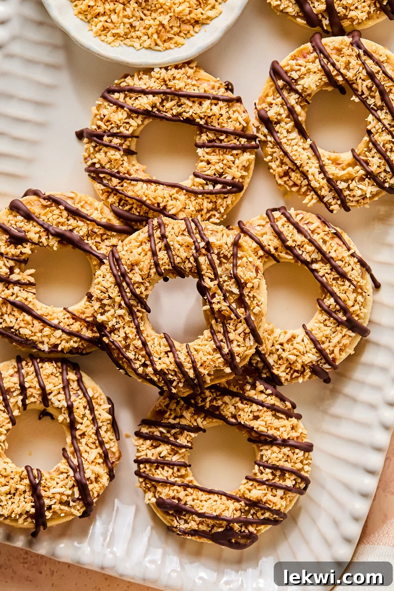A stunning close-up of Samoa cookies arranged on a platter, with a bowl of toasted shredded coconut visible in the background, highlighting the texture and detail.