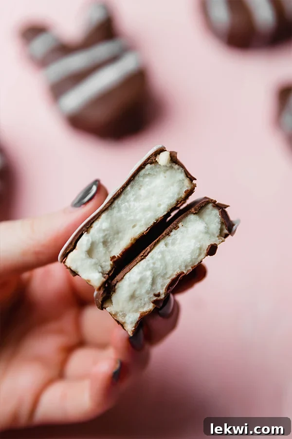 A hand gently folds a homemade marshmallow Easter egg in half, showcasing its soft, fluffy interior against a soft pink backdrop.
