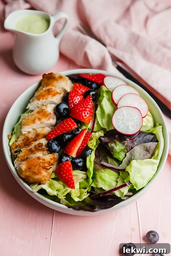 A beautiful white bowl overflowing with golden, crispy chicken slices, bright red berries, thinly sliced radishes, and mixed green lettuce, all artfully arranged to create an inviting crispy chicken salad.