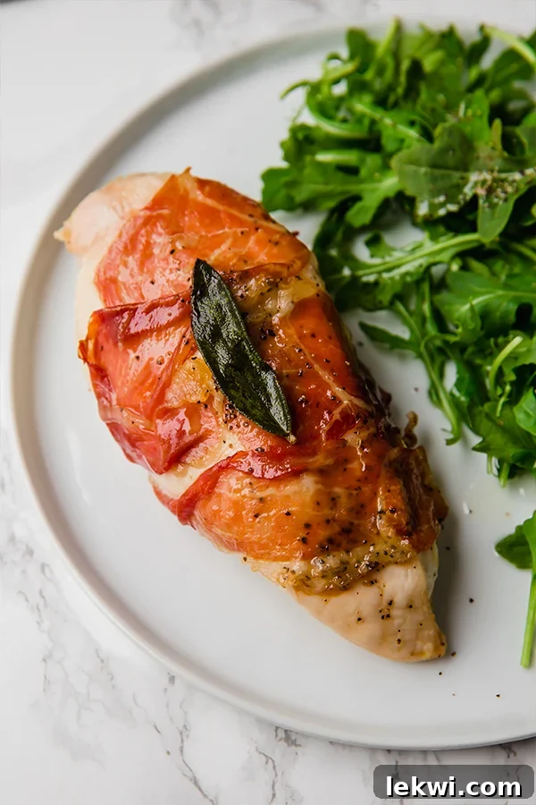 A white plate with a fresh arugula salad positioned next to a beautifully baked piece of prosciutto-wrapped chicken, garnished with herbs.