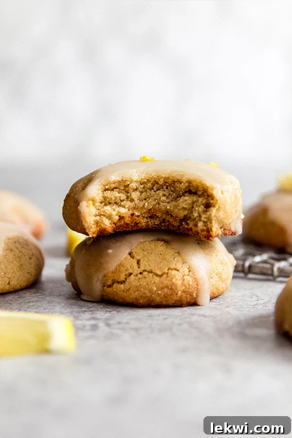 A stack of golden paleo lemon cookies, some with a white glaze, on a white plate with fresh lemon slices.