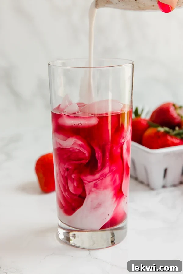 A tall glass filled with a vibrant pink drink, garnished with fresh strawberries, with more berries in the background.