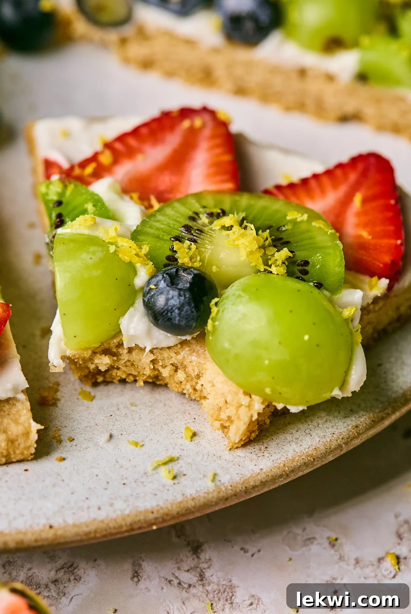 A close-up of a single slice of sugar cookie fruit pizza on a white plate, with a small bite taken out, showing the layers of cookie, frosting, and fresh fruit.