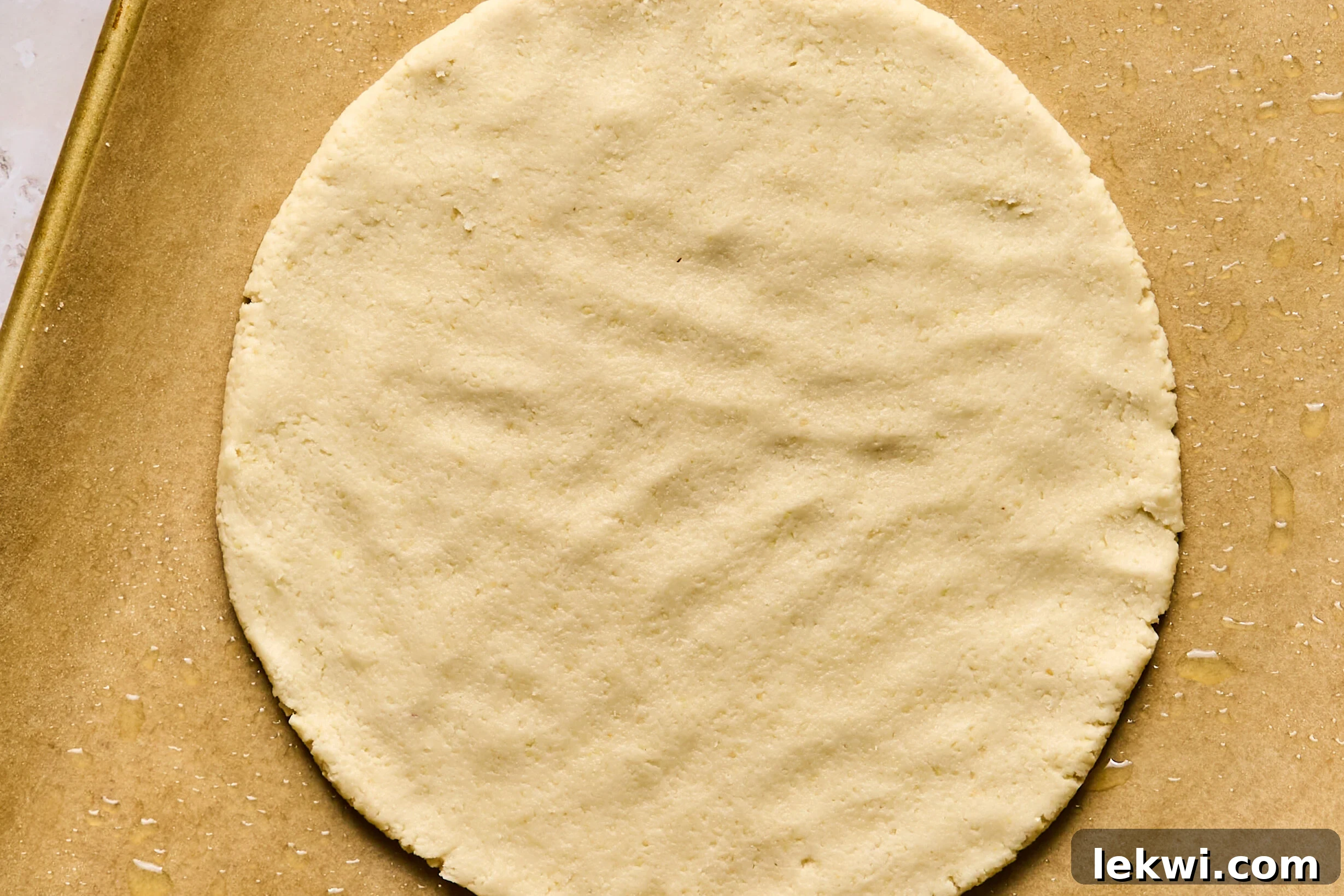 A perfectly round, unbaked sugar cookie pizza crust neatly spread on a greased baking sheet, awaiting its turn in the oven.