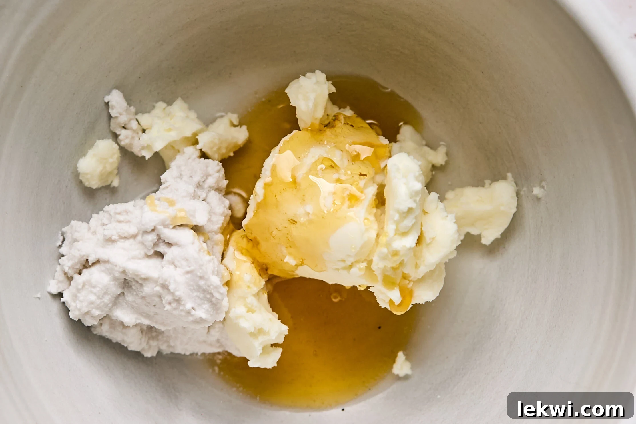 A white ceramic bowl containing separate dollops of palm shortening, rich coconut cream, and golden honey, ready to be combined for the creamy frosting.