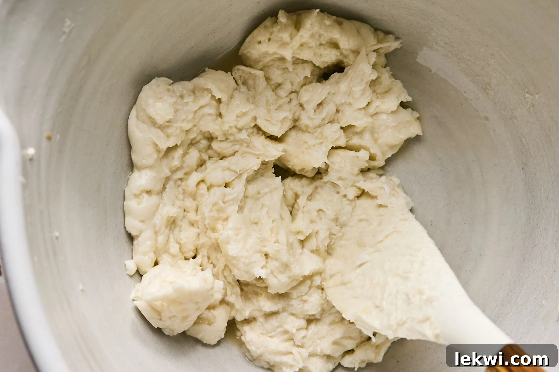 A white spatula stirring the creamy, off-white frosting mixture in a white ceramic bowl, ensuring all ingredients are thoroughly blended to a smooth consistency.