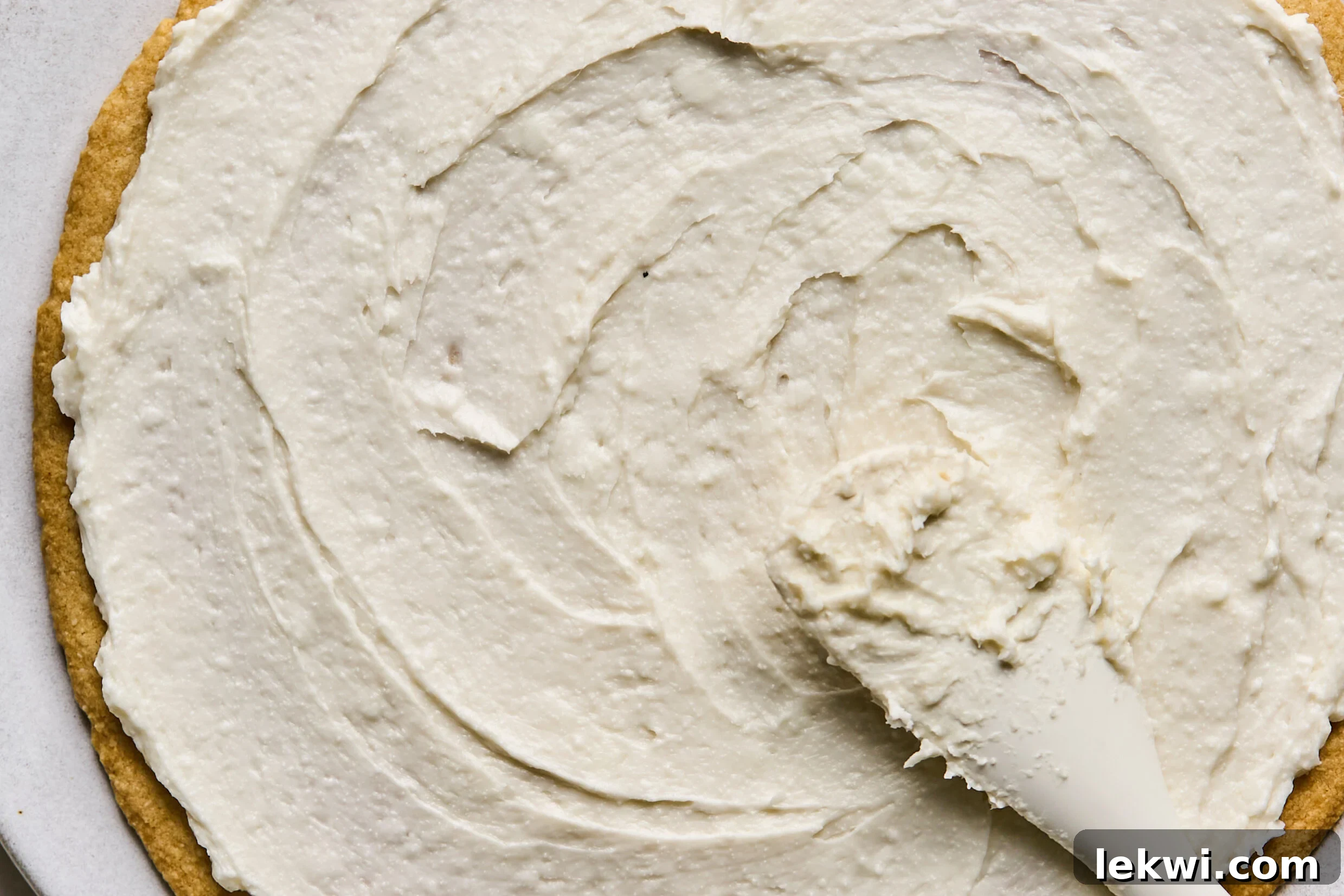 A spatula gently spreading the creamy, white frosting evenly across the golden-brown sugar cookie crust, preparing the base for the fruit toppings.