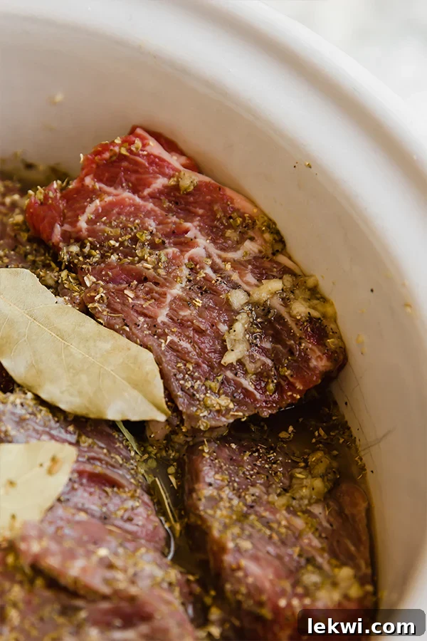 Slow cooker filled with chuck roast pieces, onions, garlic, and spices, before liquid is added.