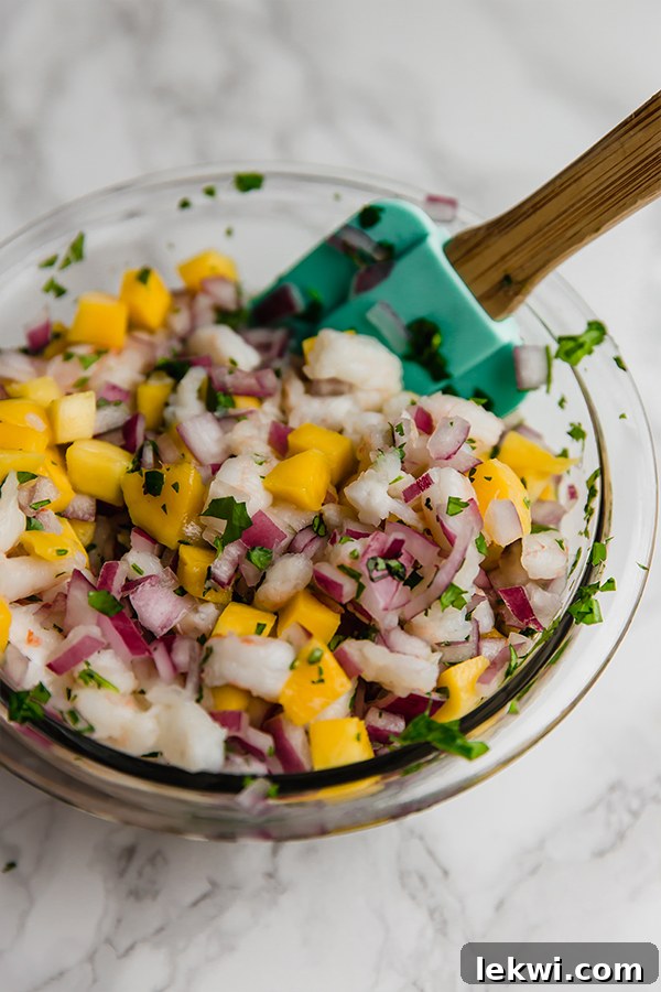 A glass bowl of vibrant mango shrimp ceviche being gently mixed with a spatula, showcasing its fresh ingredients.