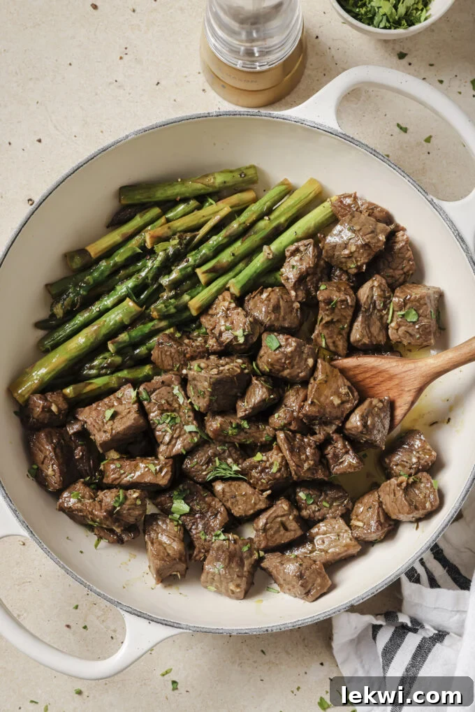 Garlic and balsamic steak bites with asparagus topped with parsley in a white pan, with a wooden spoon. 