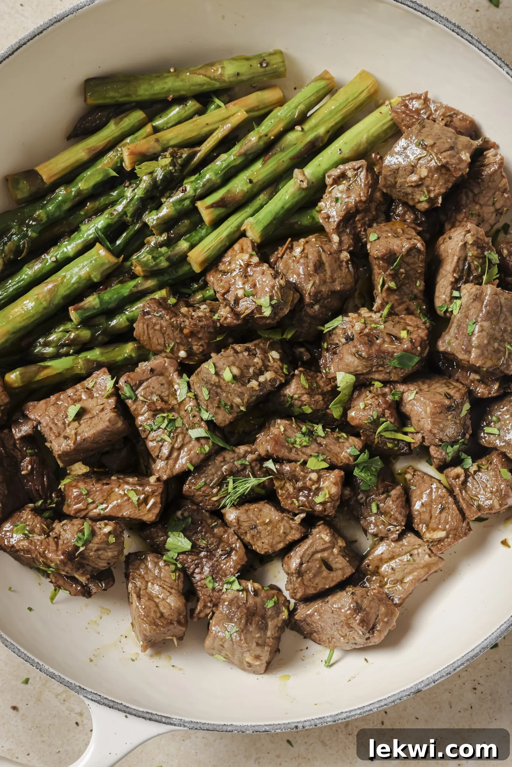 Garlic and balsamic steak bites with asparagus topped with parsley in a white pan.