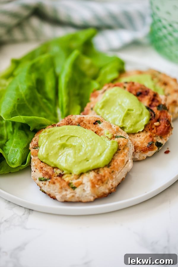 A plate with avocado chicken burgers topped with avocado and lettuce next to it.