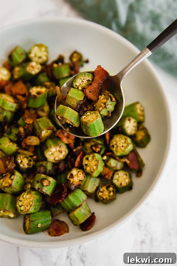 A close-up shot of a white bowl of Southern Bacon and Okra Skillet, with a silver spoon scooping out a portion, highlighting the crispy texture and vibrant colors.
