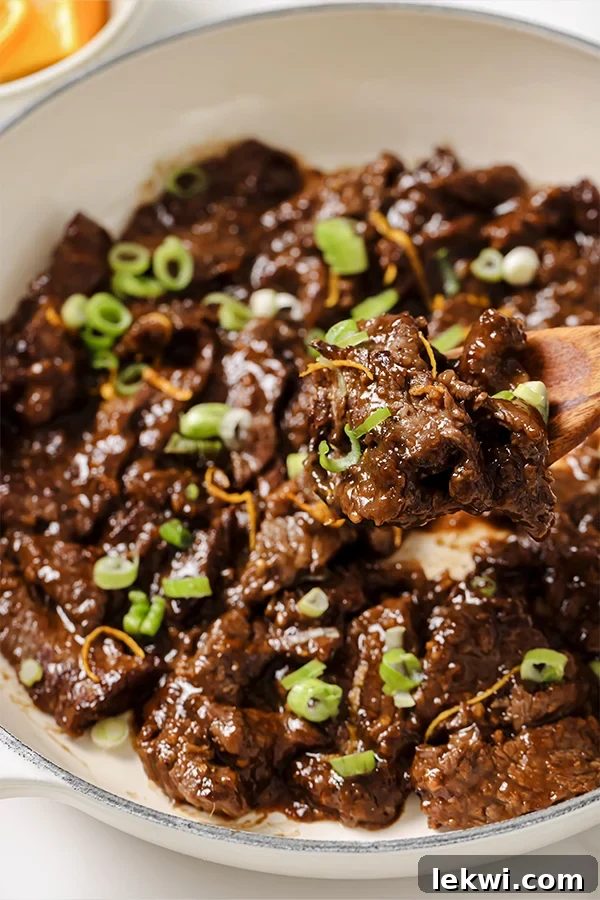 Orange beef topped with fresh green onion and orange zest, presented in a white enamel pot with a wooden spoon ready for serving.