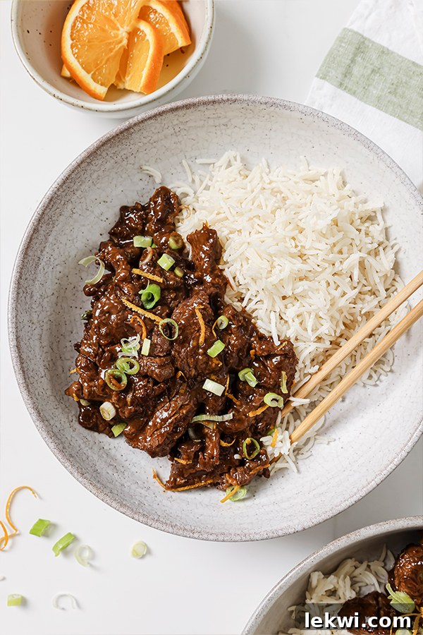 Orange beef topped with green onion and orange zest in a bowl, served alongside a portion of rice and chopsticks.