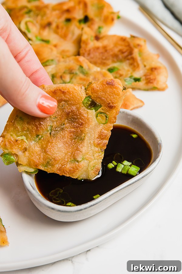 A hand dipping a quarter of a crispy gluten-free scallion pancake into a small bowl of coconut aminos dipping sauce.