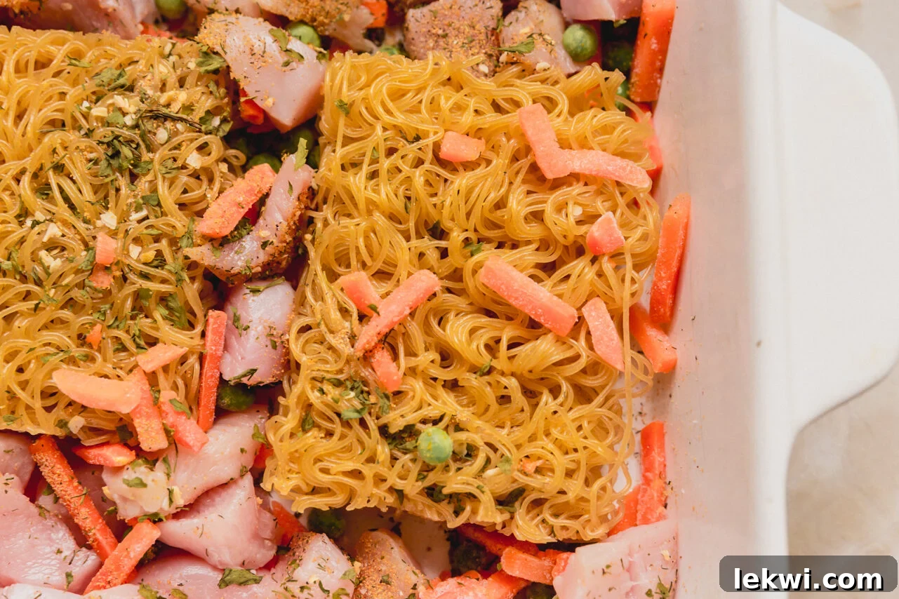 A casserole dish filled with uncooked ramen bricks, cubed chicken, and mixed frozen vegetables, seasoned and ready for liquid.