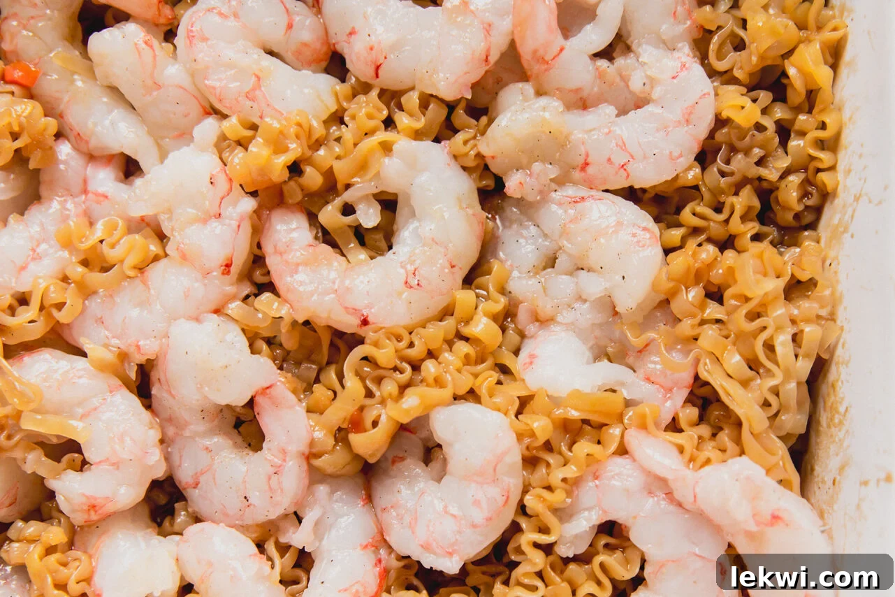 A white casserole dish with raw shrimp on top of a ramen and vegetable mixture.