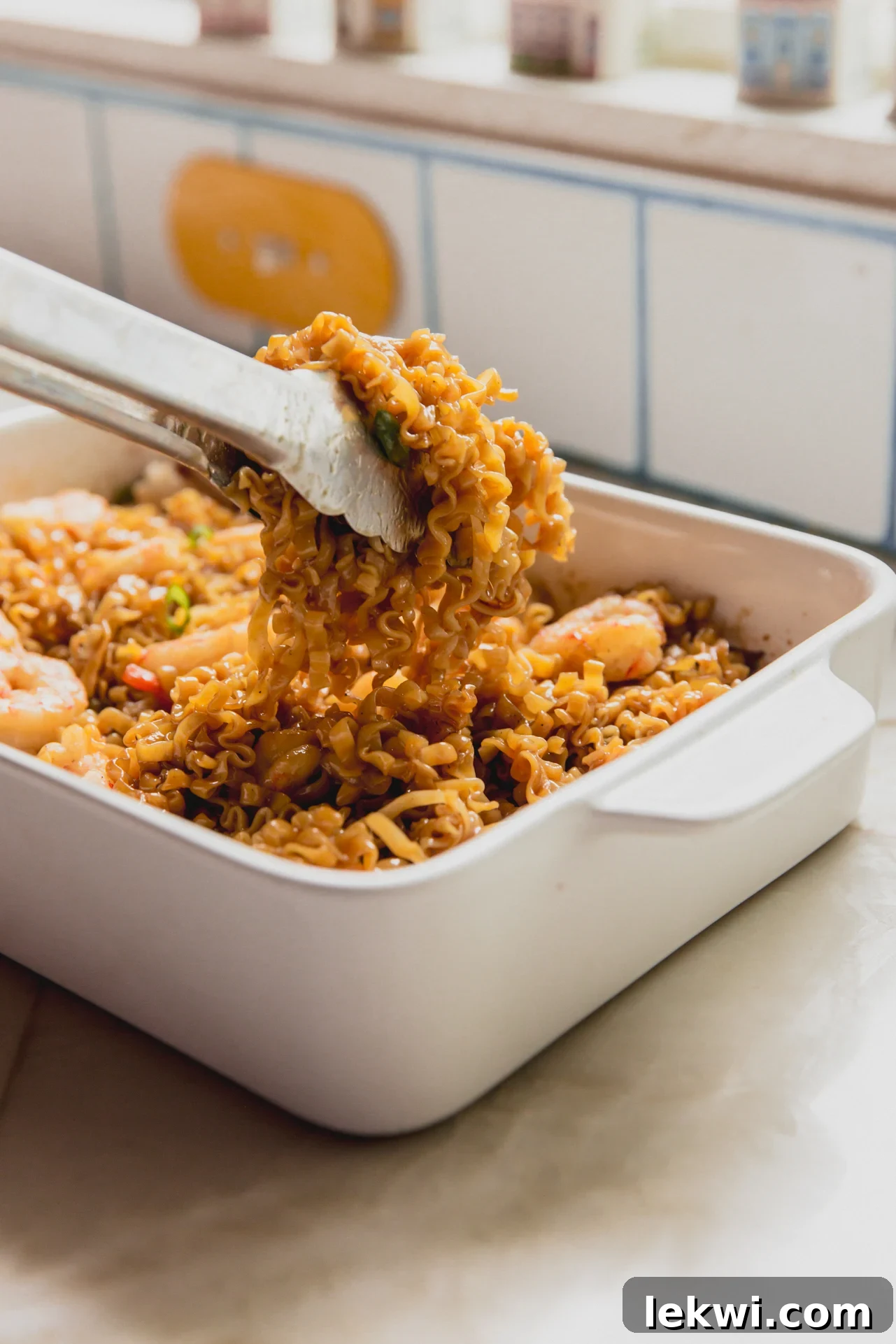 A pair of metal thongs lifting some of the shrimp ramen noodle bake out of the white ceramic dish it was baked it.